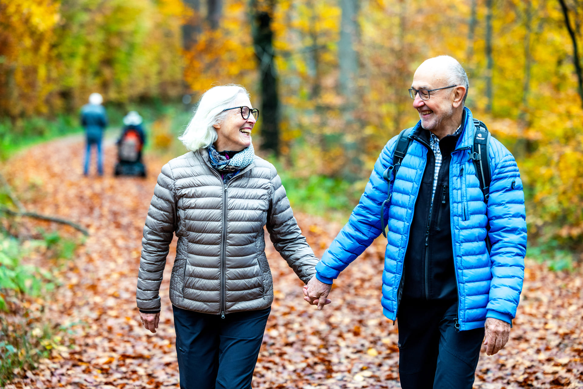 Älteres Paar läuft Händchen hlatend durch den Herbstwald. Im Hintergrund sieht man zwei weitere Personen, eine davon sitzt im E-Rolli.