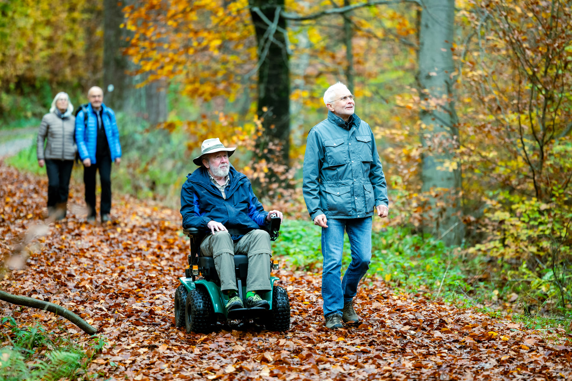 Älterer Herr im Outdoor-Rollstuhl läuft mit Begleiter durch den Herbstwald