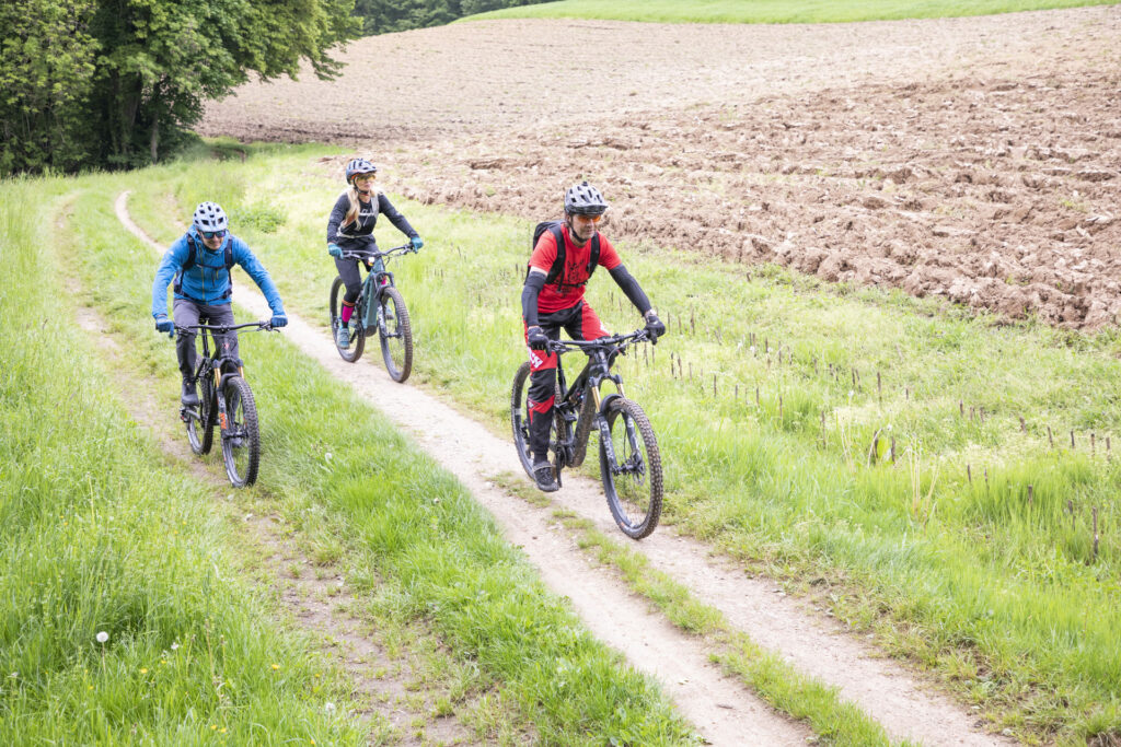 Drei Mountainbiker fahren auf einem Schotterweg an einem Feld entlang